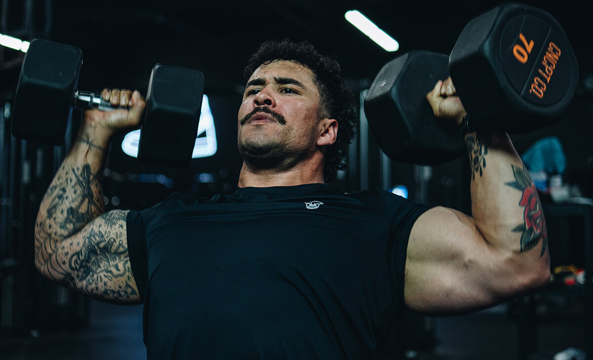Athlete Jack West performing a dumbbell shoulder press in a gym, wearing a Gymreapers Performance Short Sleeve Shirt and Performance Shorts.